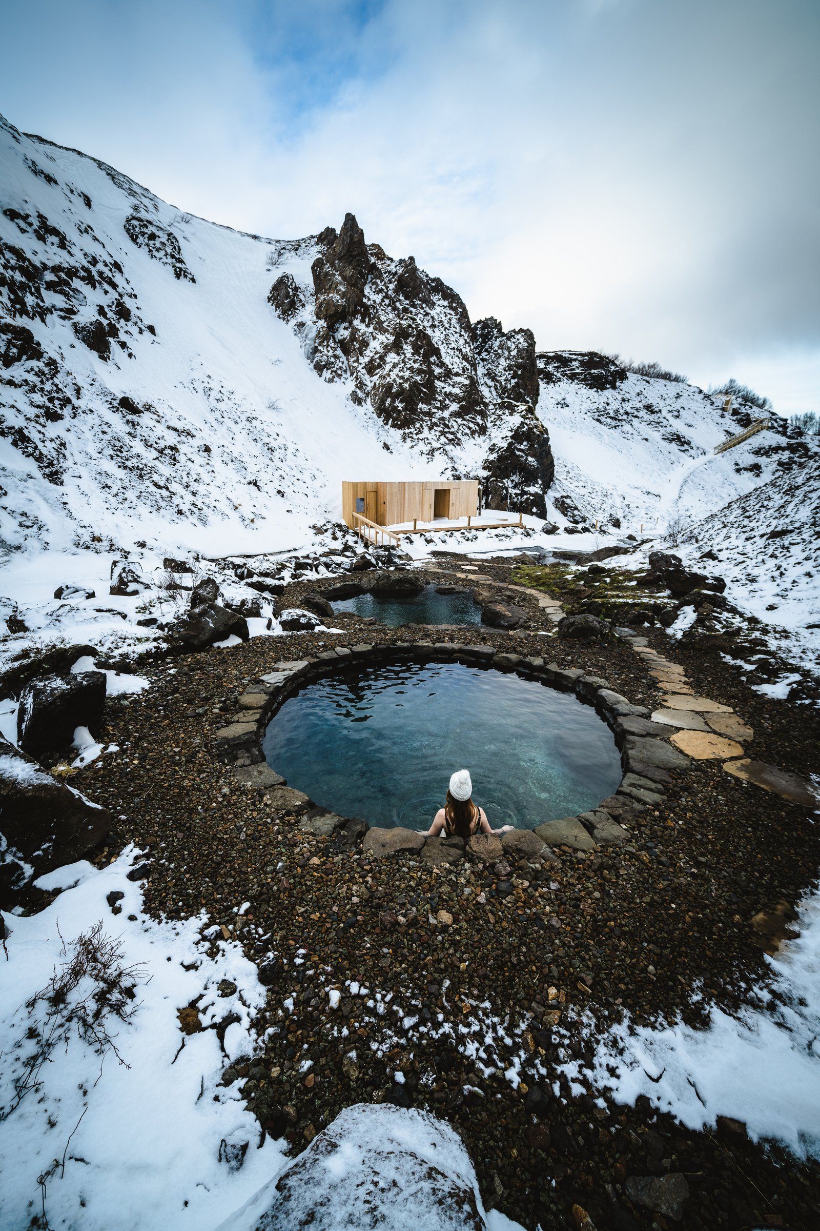 A person relaxes in a geothermal pool at the Húsafell Canyon Baths, surrounded by snow-covered hills and rugged volcanic landscapes in Iceland.