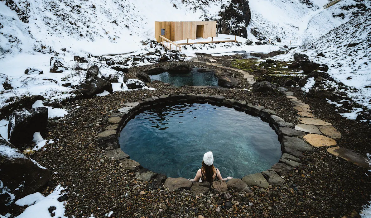 A person relaxes in a geothermal pool at the Húsafell Canyon Baths, surrounded by snow-covered hills and rugged volcanic landscapes in Iceland.