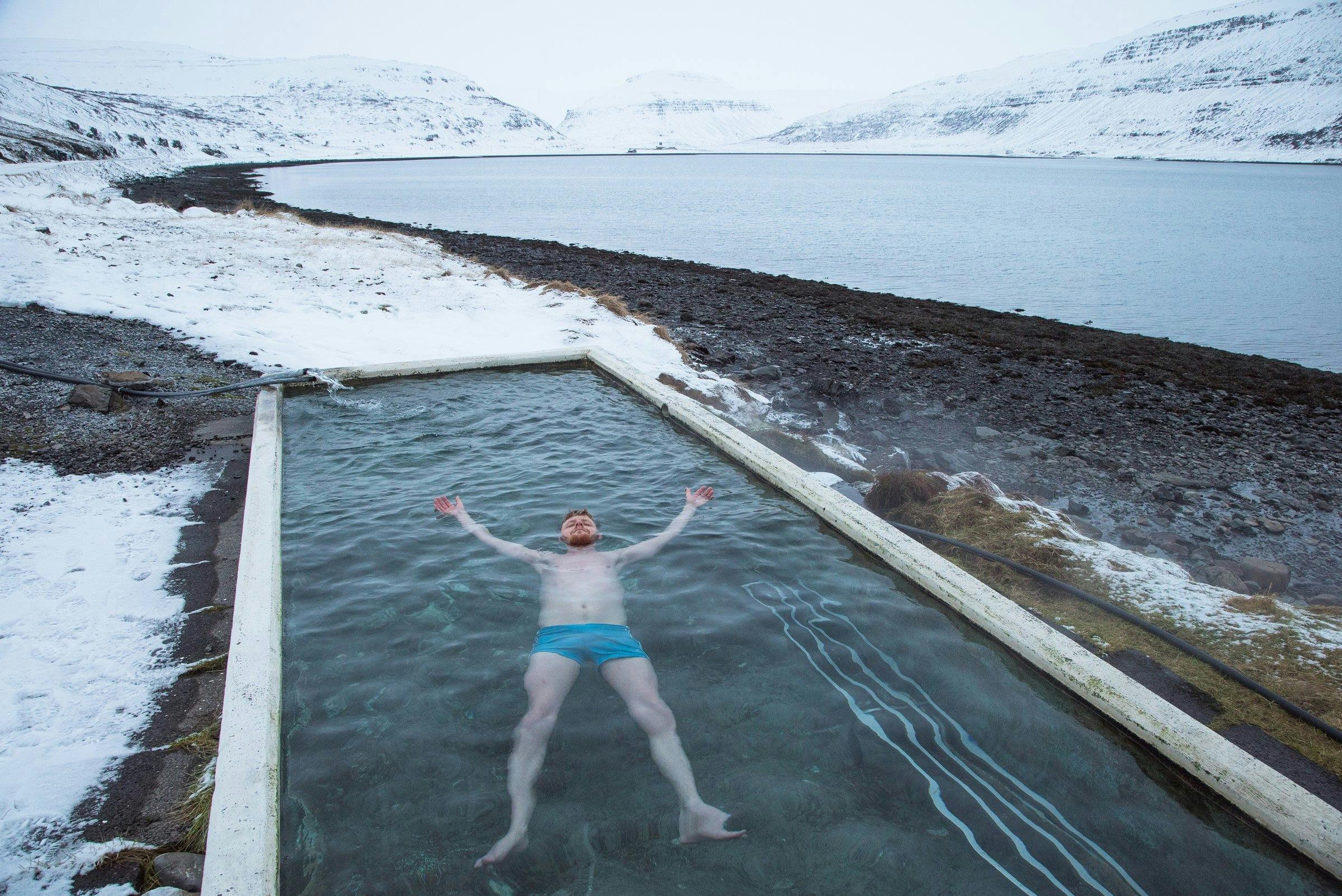 Man in blue bathing shorts lying in a hotwater pool surrounded only by snowy mountains