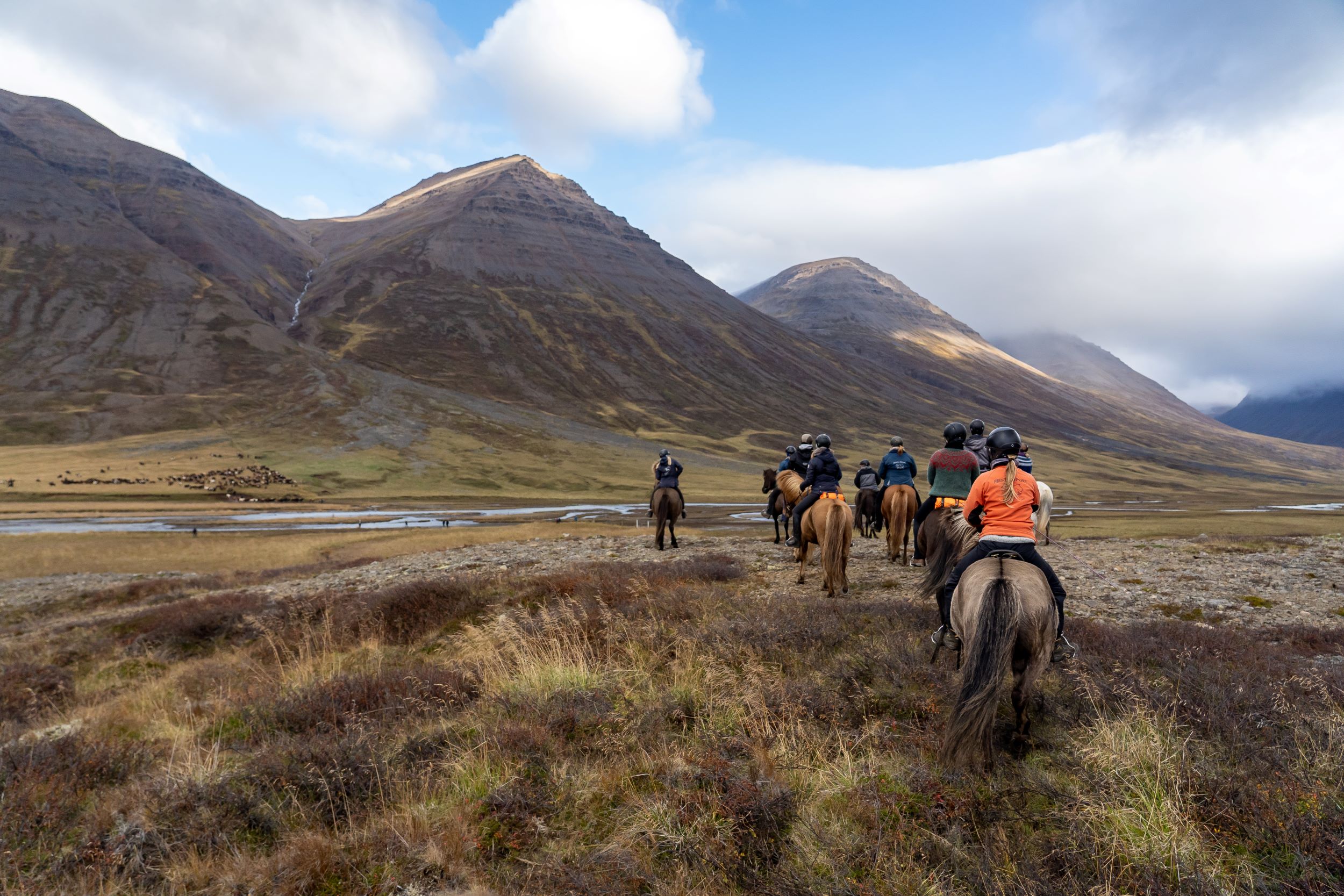 A group of riders waits for a large herd of horses to cross a river. High mountains in the backdrop.