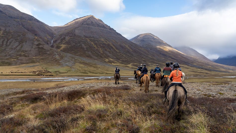 A group of riders waits for a large herd of horses to cross a river. High mountains in the backdrop.