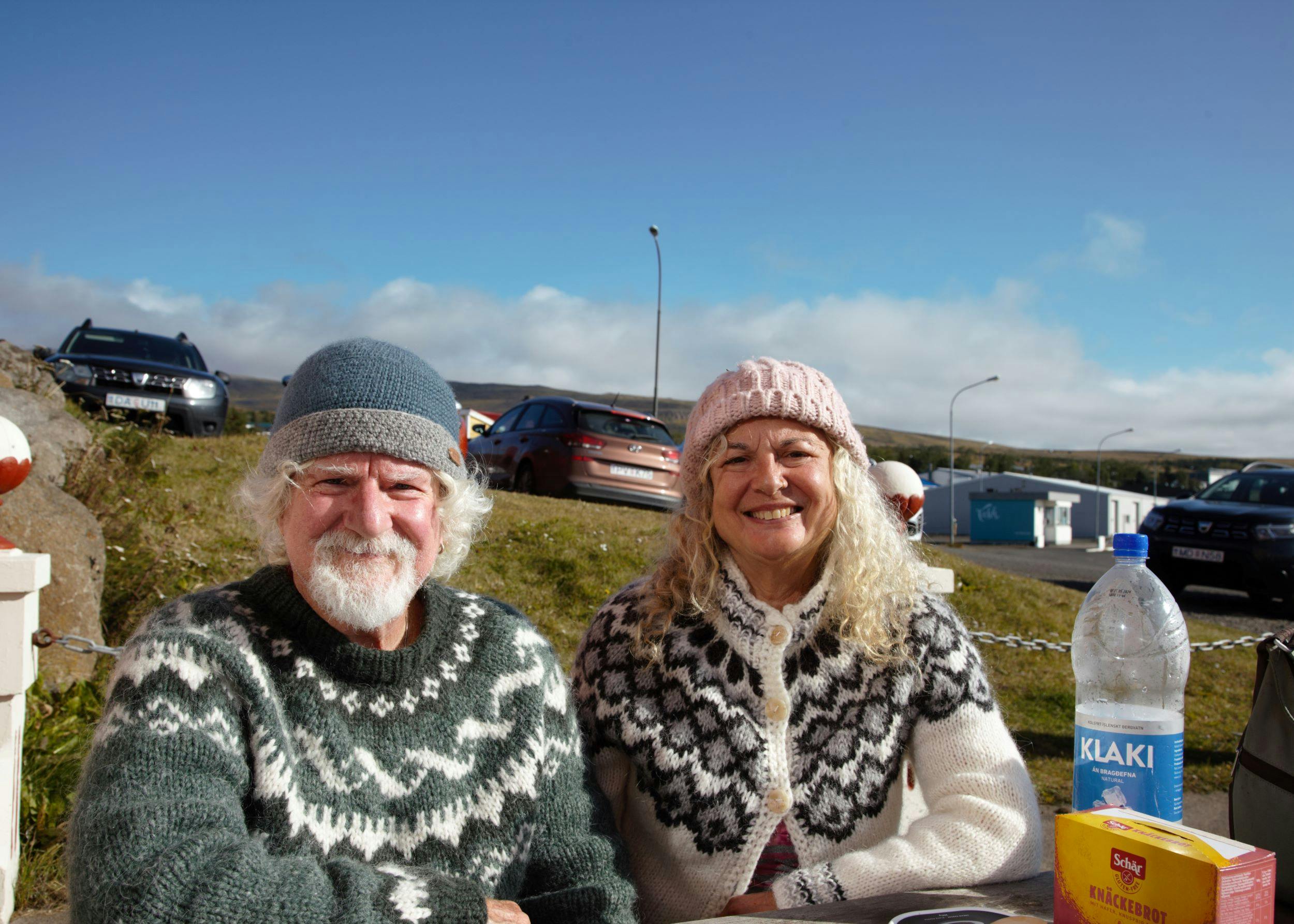 Elderly couple in handknitted Icelandic sweaters sitting on a picnic table, smiling, the sun i shining, cars in the background