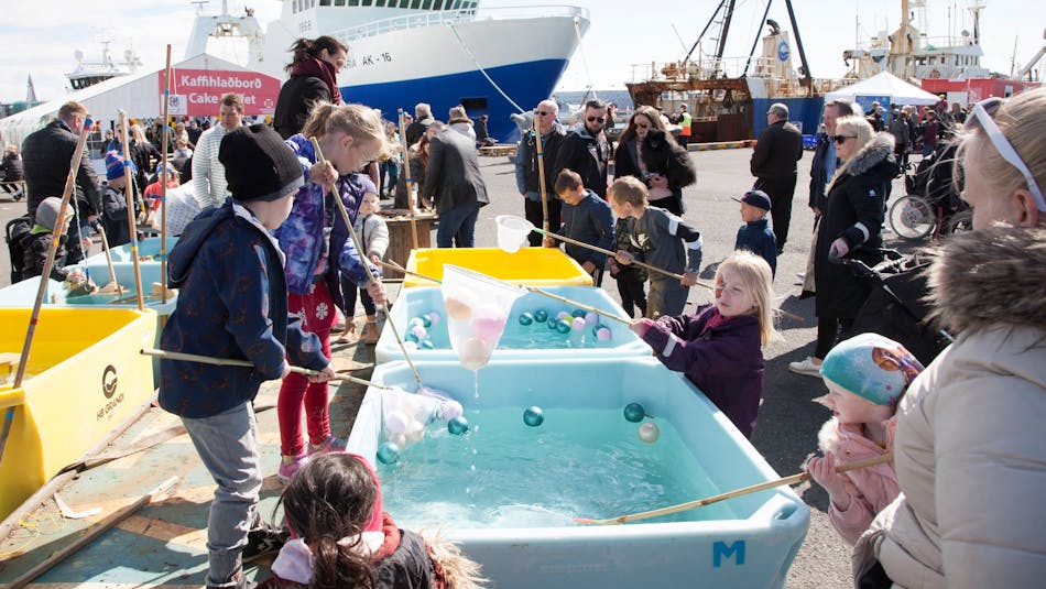 Children fishing colourful balls out of water bassins in Reykjavík harbour. Fishing vessels in the background