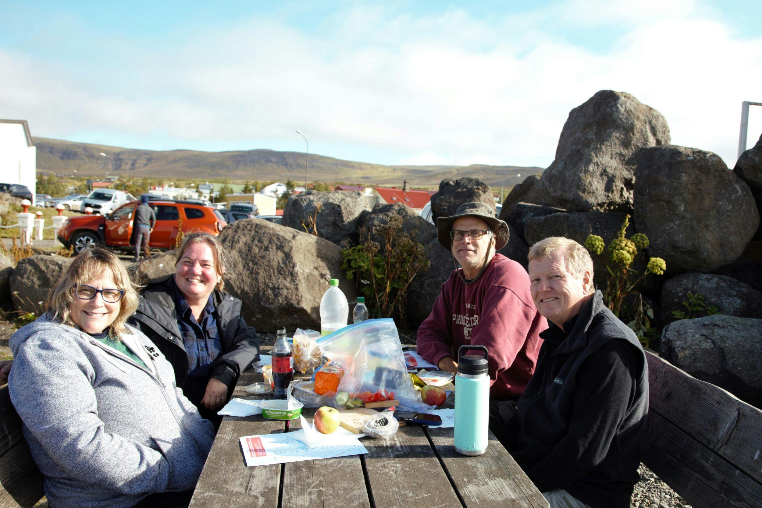 Two women and two men sitting at a picnic table having lunch, smiling into the camera. Mountains in the background