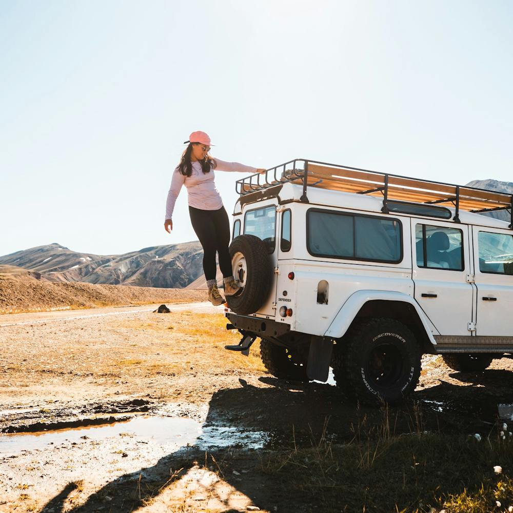 Woman standing on the spare wheel of a white jeep, parked in the Icelandic highlands. She is wearing a thin long sleeve, black pants, hiking boots and sunglasses. The sun is shining.