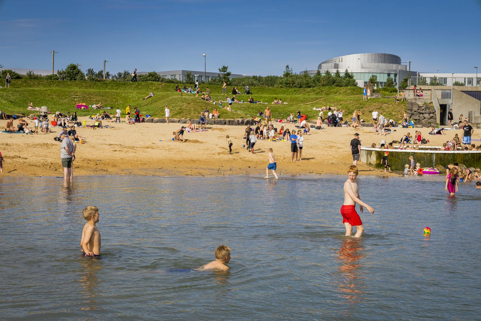 Nautholsvík beach on a summer day, children are swimming in the ocean, in the background there is a yellow sand beach with groups of people enjoying the sun