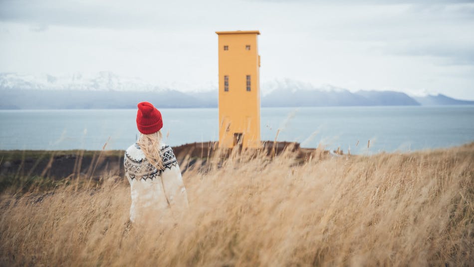 Young woman wearing a lopapeysa in front of a yellow lighthouse in North Iceland