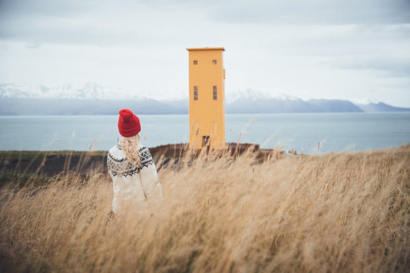 Young woman wearing a lopapeysa in front of a yellow lighthouse in North Iceland