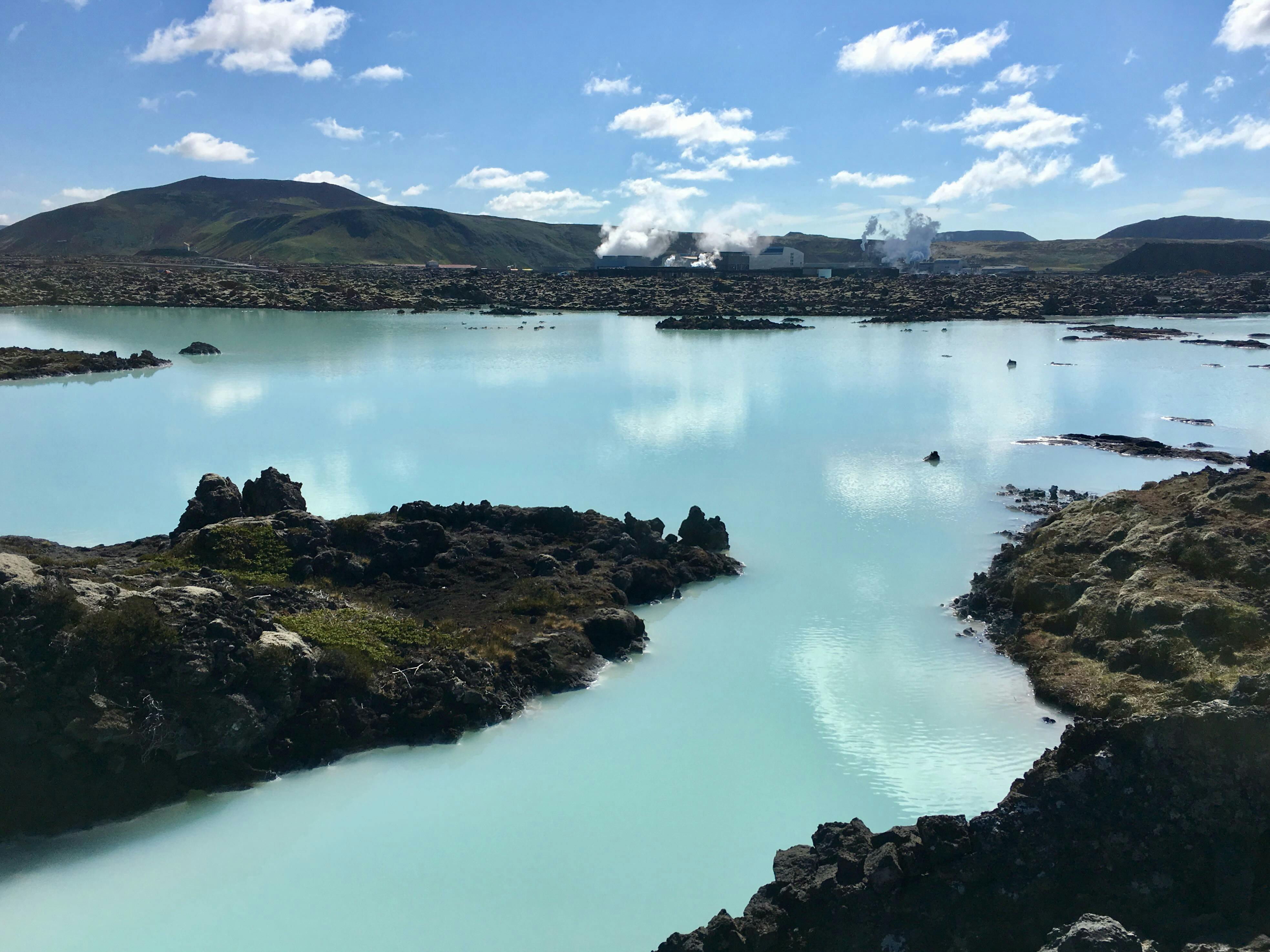 A bright blue lake in a black lava landscape and steam rising from buildings in the back