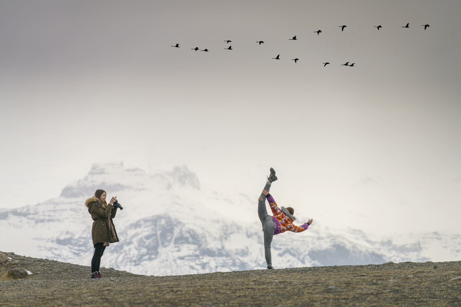 A traveler poses playfully while another takes a photo against a backdrop of snow-covered mountains in East Iceland, as a flock of birds flies overhead.
