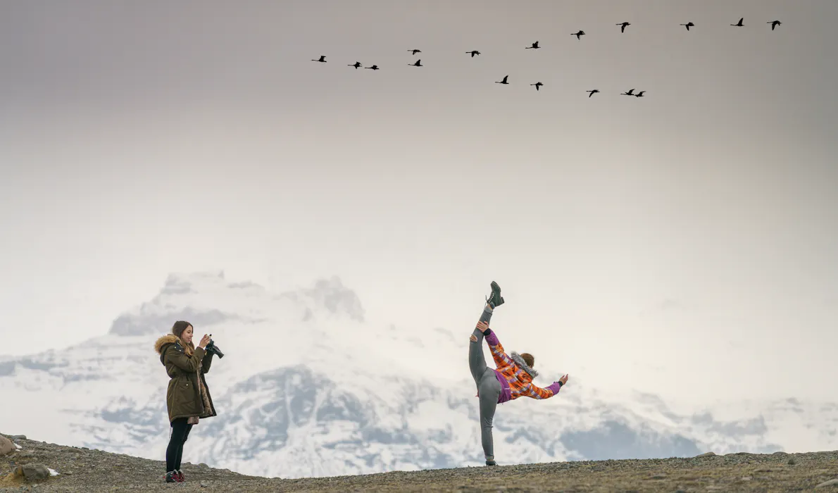A traveler poses playfully while another takes a photo against a backdrop of snow-covered mountains in East Iceland, as a flock of birds flies overhead.