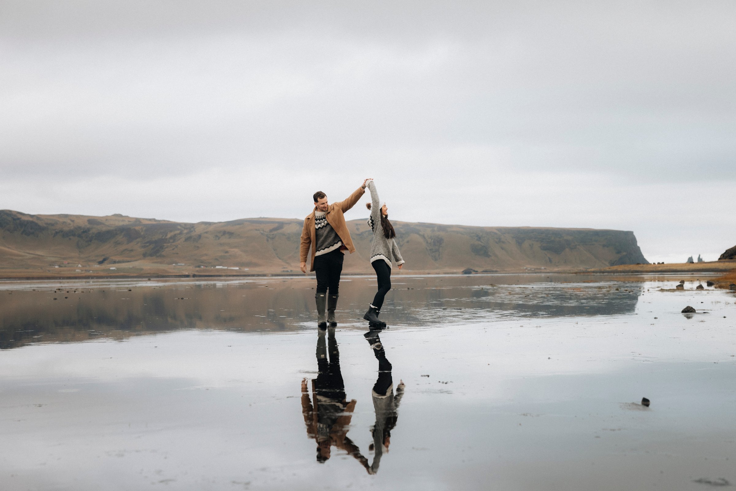 A couple holds hands and twirls across the reflective black sands of Reynisfjara beach, with dramatic sea cliffs and sea stacks in the background.