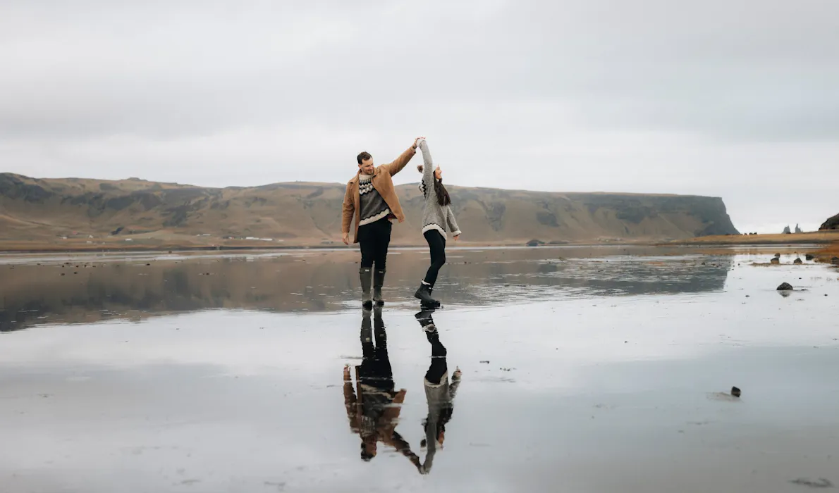 A couple holds hands and twirls across the reflective black sands of Reynisfjara beach, with dramatic sea cliffs and sea stacks in the background.