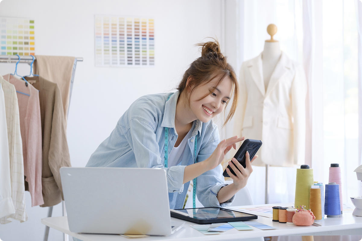 Smiling merchants using a smartphone in her studio surrounded by fabric swatches and a dress form, representing a small business owner accepting online payments with viva.com