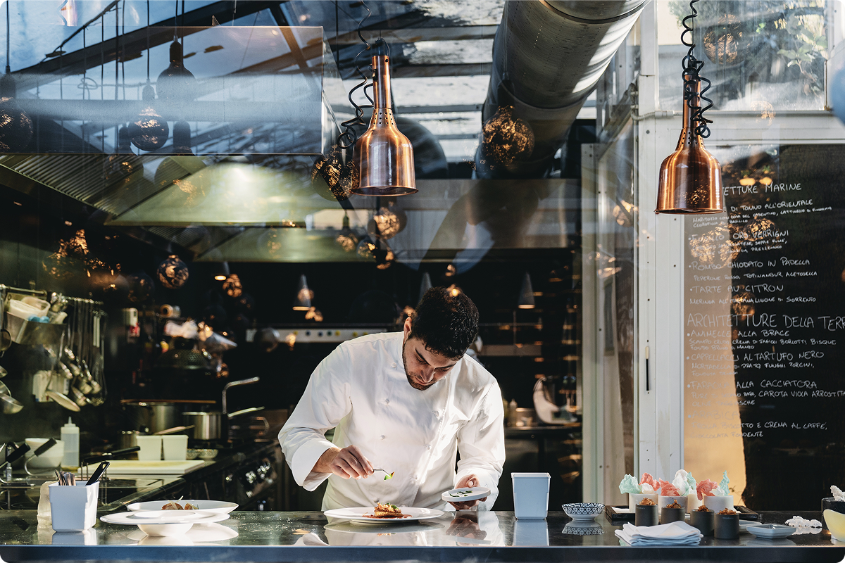A chef in a white jacket carefully plating a dish at the pass of a professional open kitchen, with copper heat lamps overhead and a handwritten menu visible in the background, representing the hospitality and HORECA sector served by Viva.com payment solutions