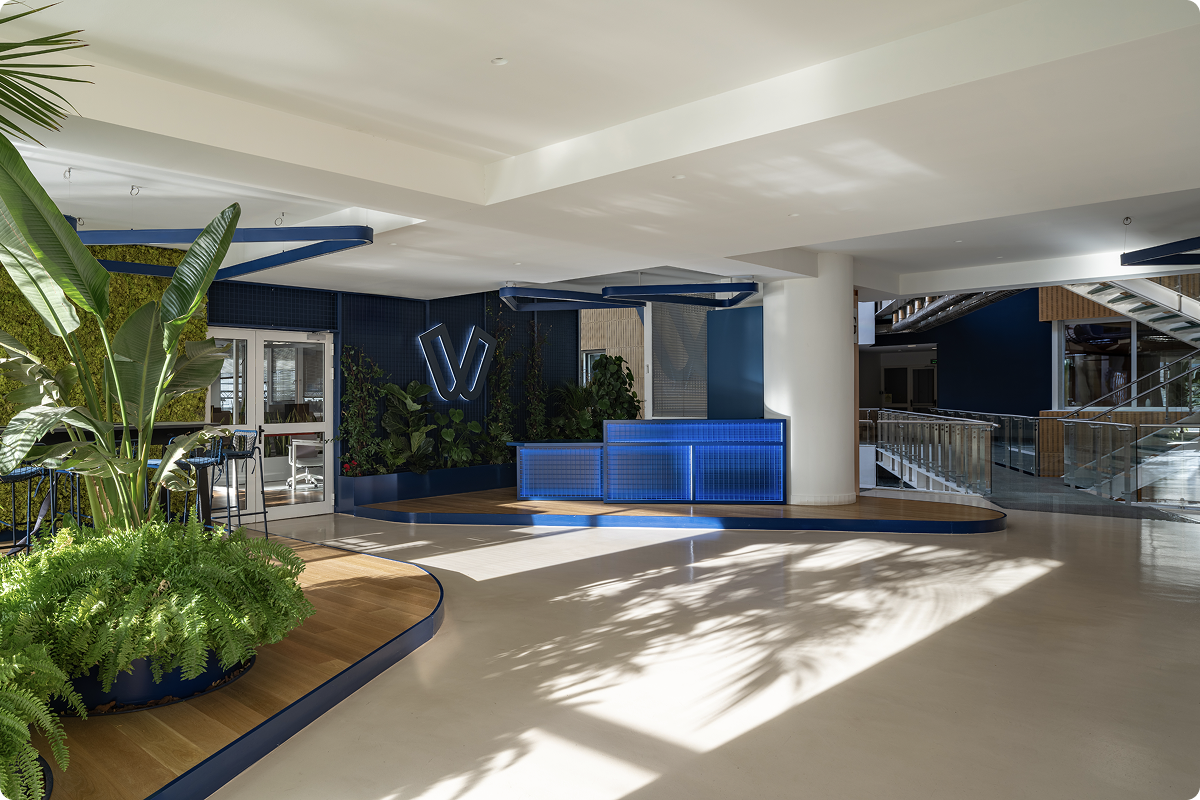 The reception area of the Viva.com headquarters featuring a glowing blue front desk, an illuminated Viva.com logo on a dark navy wall, lush indoor plants, and an open staircase in a modern, sunlit office space