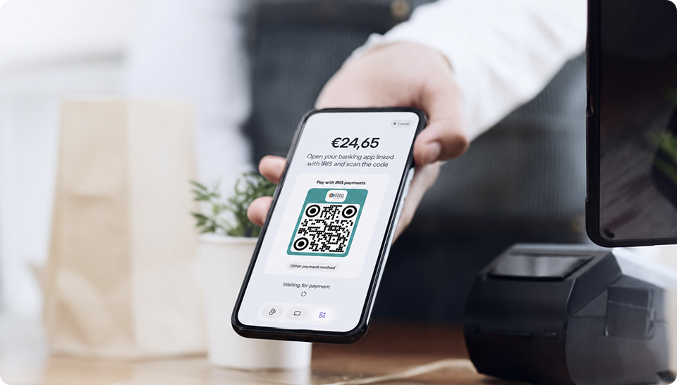 A merchant holding a smartphone displaying a Viva.com checkout screen with an IRIS QR code payment request for €24.65, next to a POS terminal on a café counter, prompting the customer to scan the code using their IRIS-linked banking app.