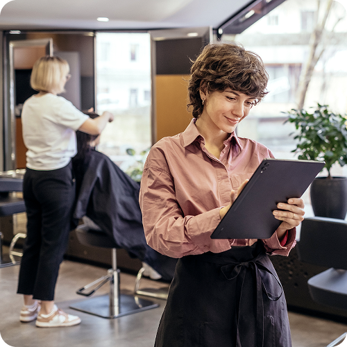 Hair salon owner smiling while using a tablet to manage her business, with a hairdresser styling a client's hair in the background
