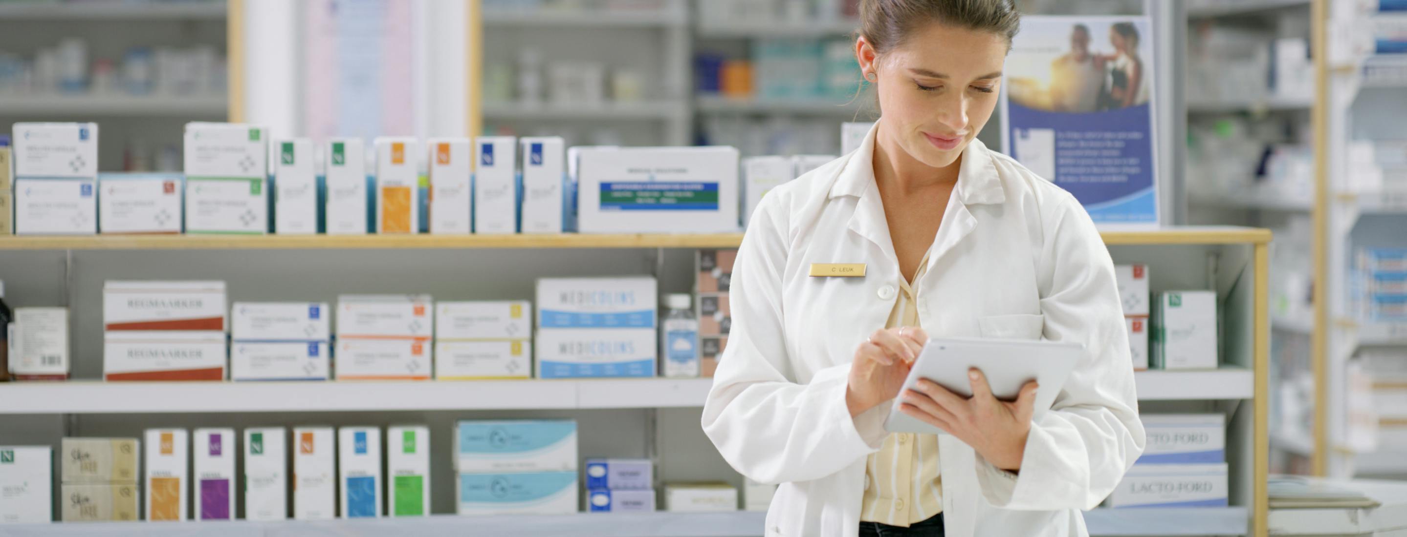 Pharmacist in white coat using a digital tablet behind the counter in a modern pharmacy, managing prescriptions and digital transactions.
