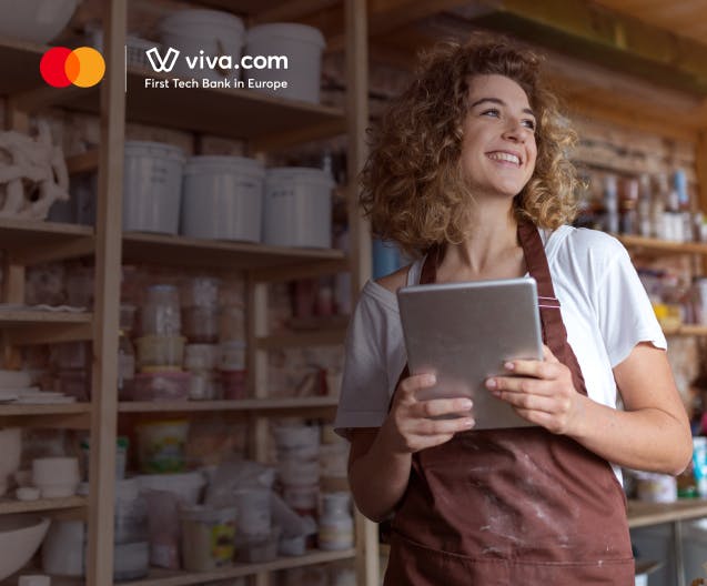 Smiling small business owner holding a tablet in a pottery art studio with shelves of clay supplies and tools