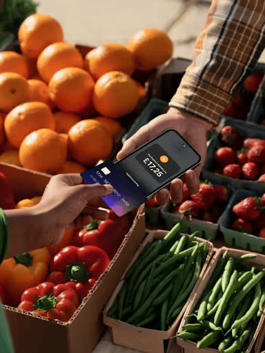 Customer making contactless payment using Tap to Pay on iPhone at farmers market stand with fresh oranges, tomatoes, peppers, and green beans