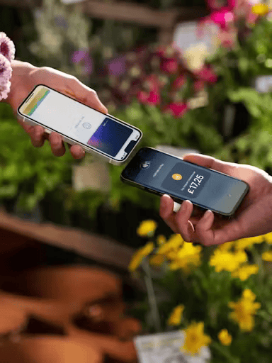 Two people completing Tap to Pay on iPhone transaction in garden setting - merchant phone showing £17.25 payment amount and customer phone with digital wallet approaching for contactless payment