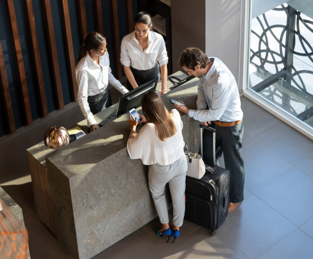 Two hotel guests standing at a modern reception desk, paying with a smartphone while two receptionists assist them, with a suitcase beside the counter.