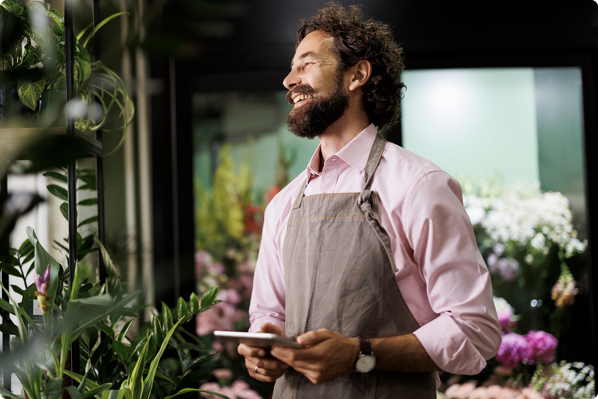A smiling small business owner wearing an apron holds a tablet in a flower shop, representing a Viva.com merchant accessing cash advance financing to grow their business