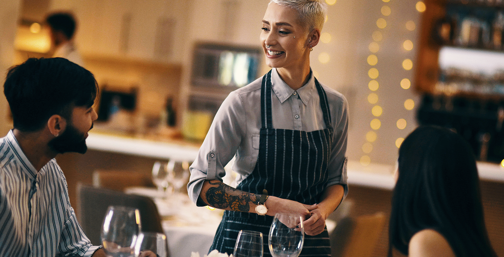 Smiling waitress in an apron greeting customers at a restaurant table, representing excellent hospitality service during the festive season