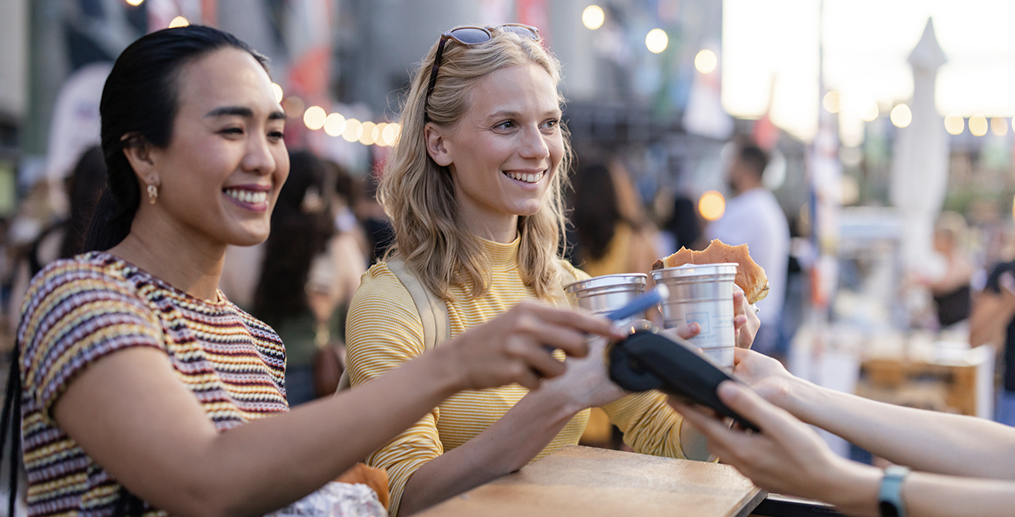 Two smiling women paying contactlessly at an outdoor street food market counter, illustrating seamless international payment acceptance and Dynamic Currency Conversion for merchants