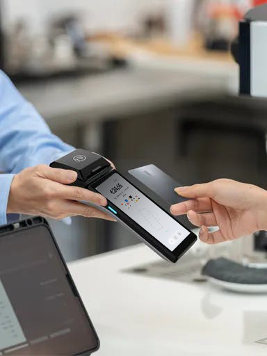 Customer making a contactless card payment using a card on an Android POS device running the viva.com Terminal app, with the merchant holding the payment terminal at a retail checkout counter.