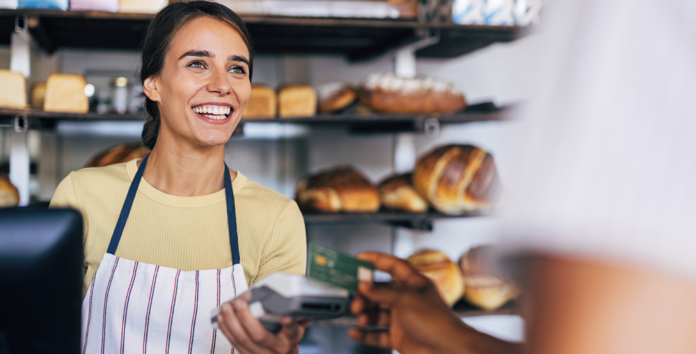 Smiling bakery owner holding a card terminal while a customer pays by card, with shelves of fresh bread in the background, illustrating card payment acceptance for small businesses
