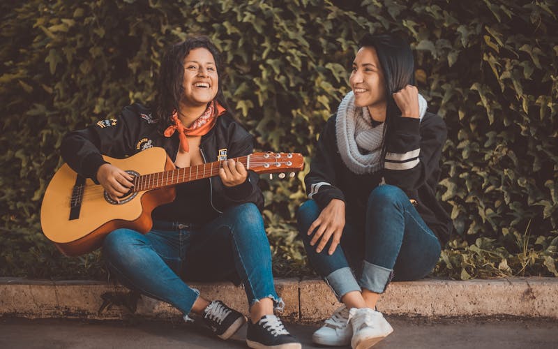 Two women sat together in a street playing the guitar and laughing