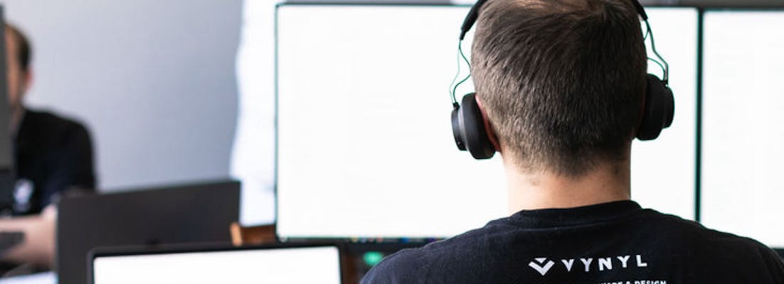 man sitting at computer desk looking at computer monitor wearing headphones