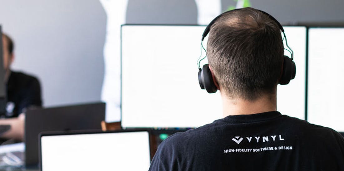 man sitting at computer desk looking at computer monitor wearing headphones