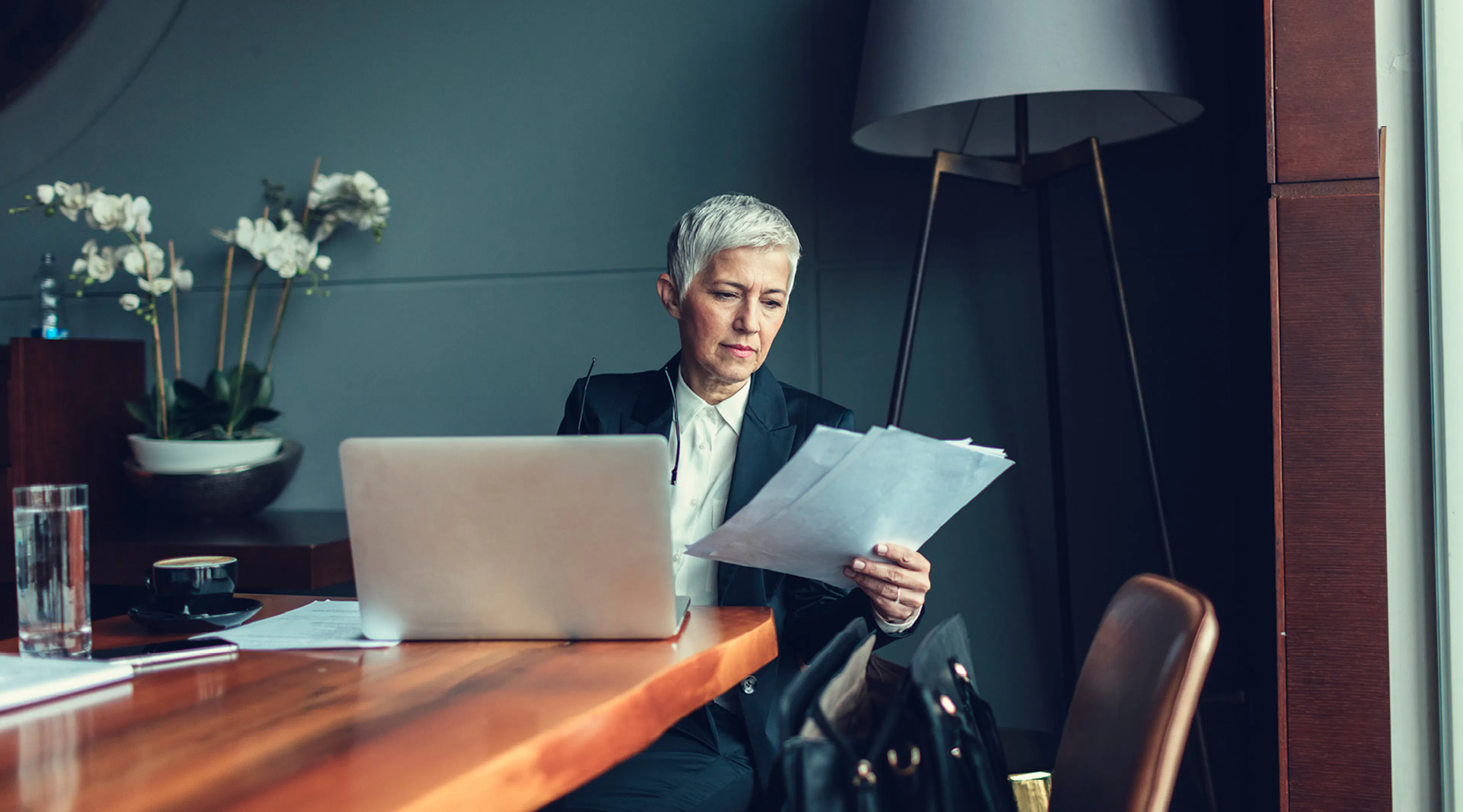 This is an image of a lady with short grey hair, wearing a dark suit and white shirt, seated at a wooden desk looking at papers, there is a dark green wall behind her and white flowers on the desk. it is an atmospheric image.