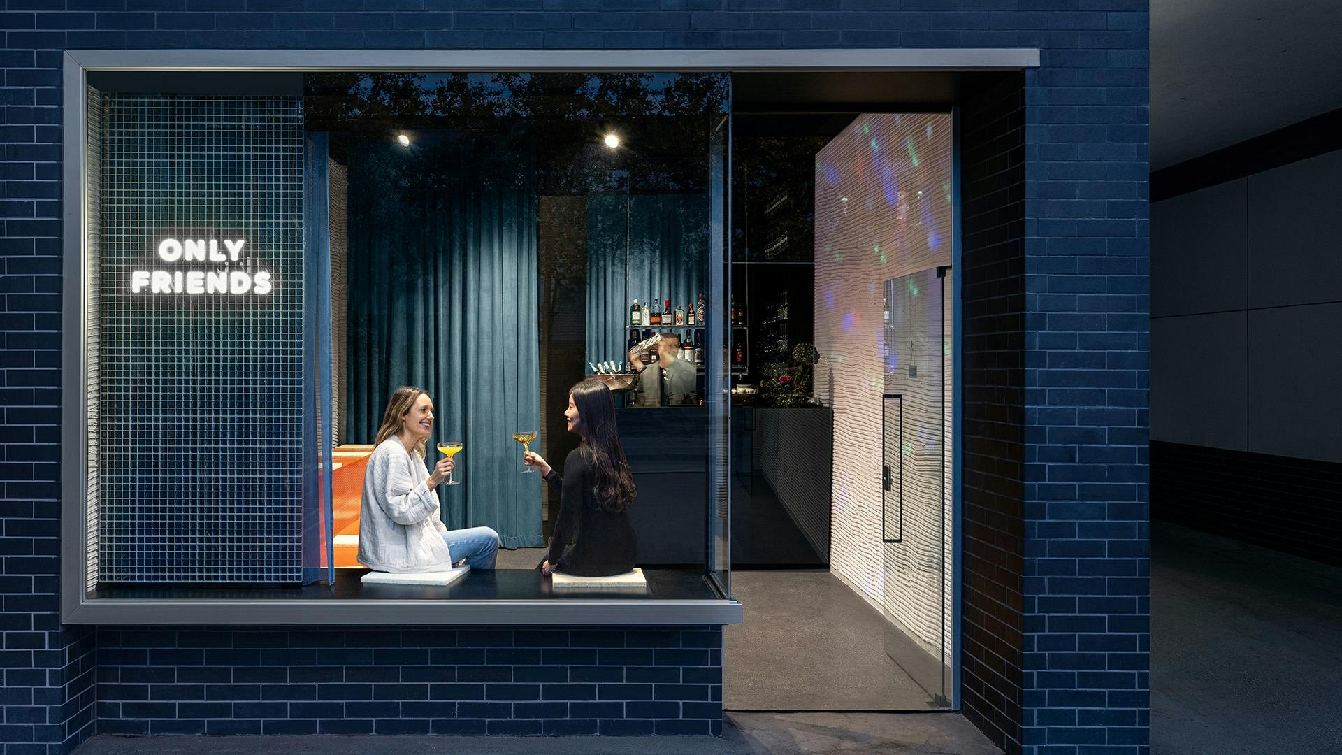 Zwei Frauen stoßen mit Cocktails in der Bar „Only Friends“ am Fenster an.