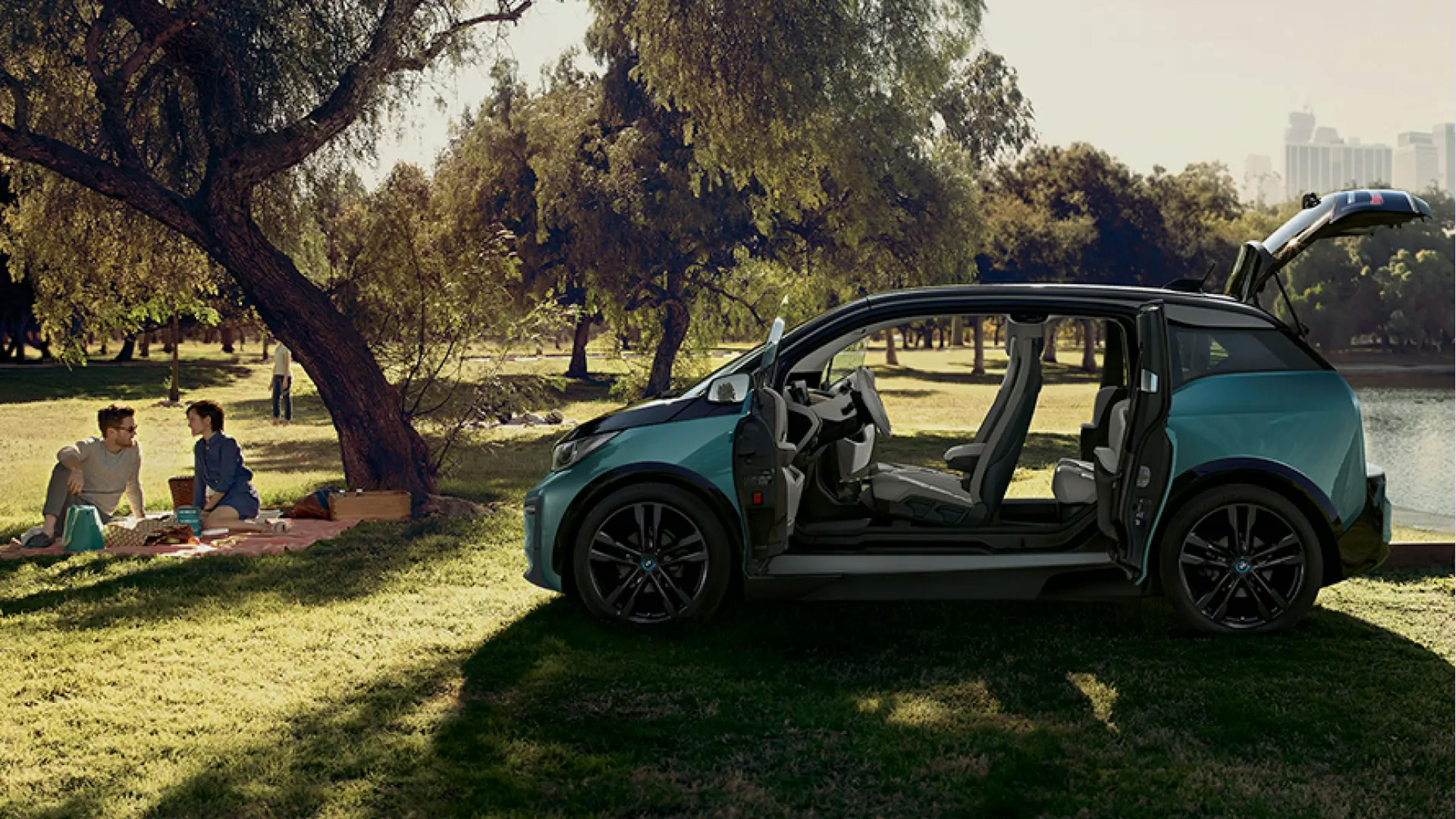 BMW i3 mit offenen Türen im Park, daneben picknickendes Paar unter einem Baum