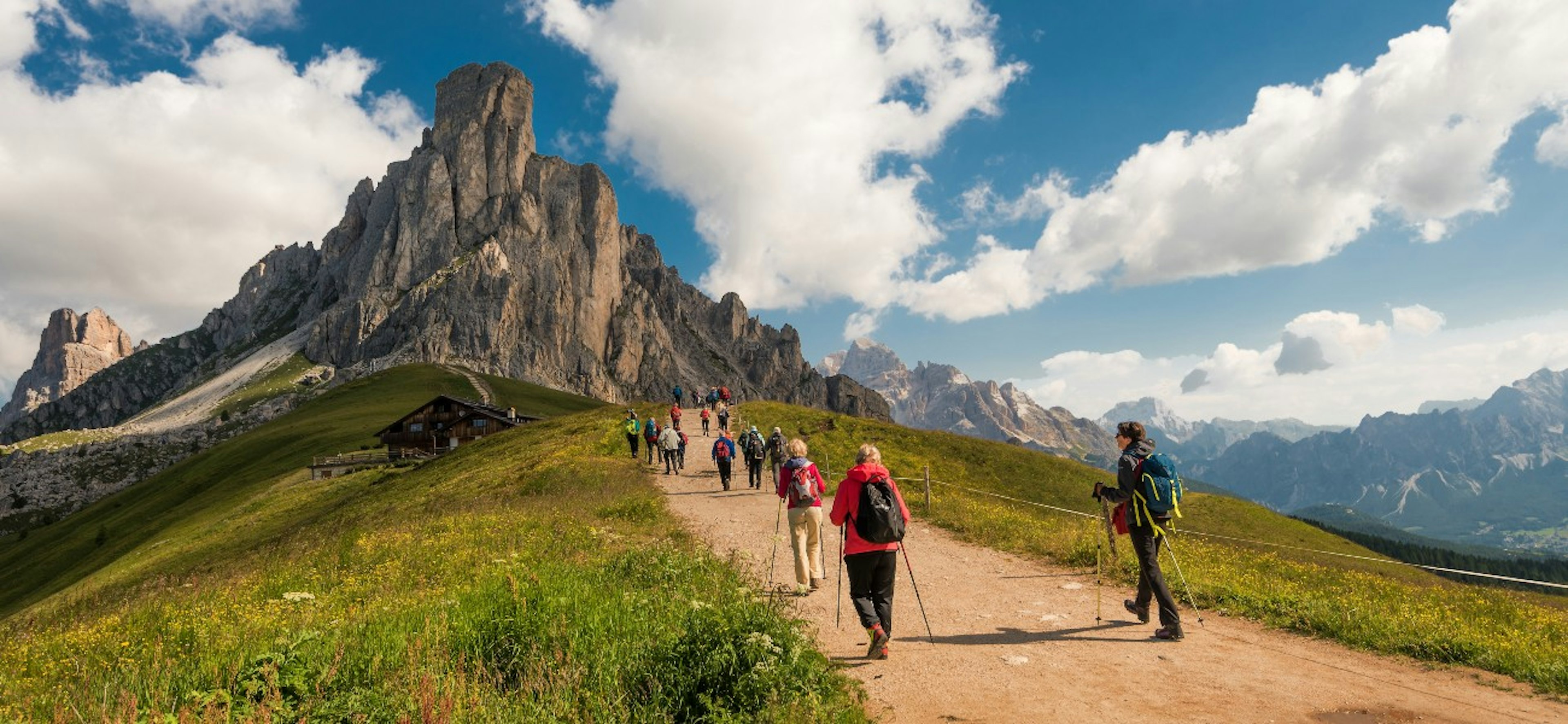 beautiful view of the Dolomites with hikers on path and grass in the foreground and hut, mountains and blue sky with clouds in the background taken on a sunny summer day