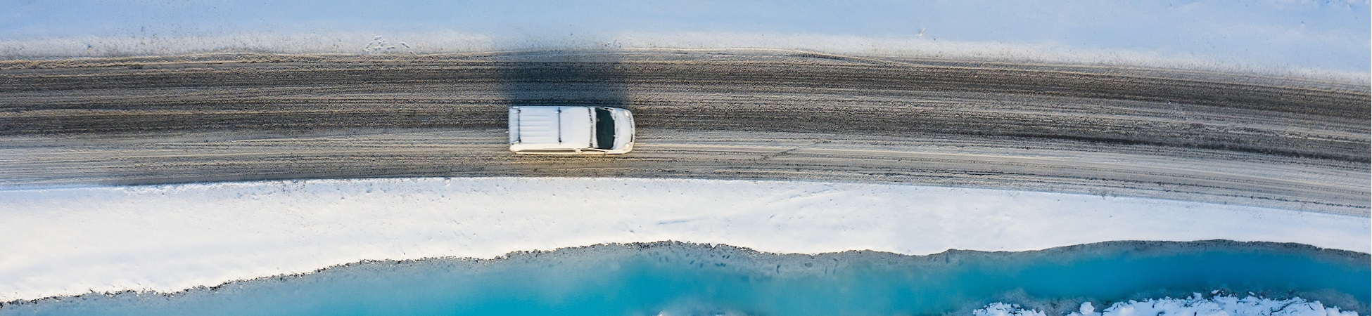 A drone shot of a car driving on a lonely road next to a blue lagoon in a snowy lava landscape