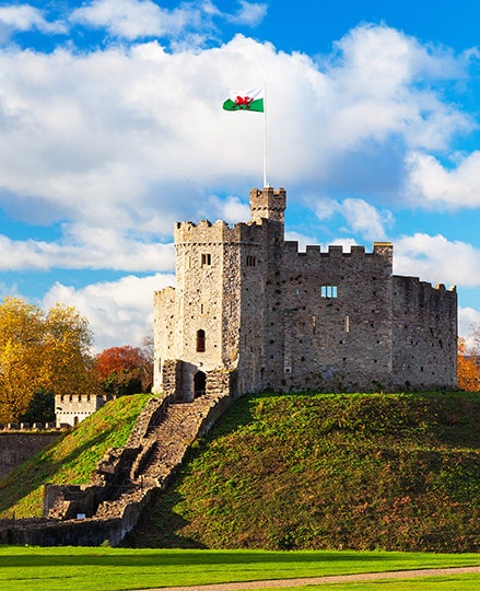 A castle on a hill with the Wales flag on top and blue sky and some cloud in the background.