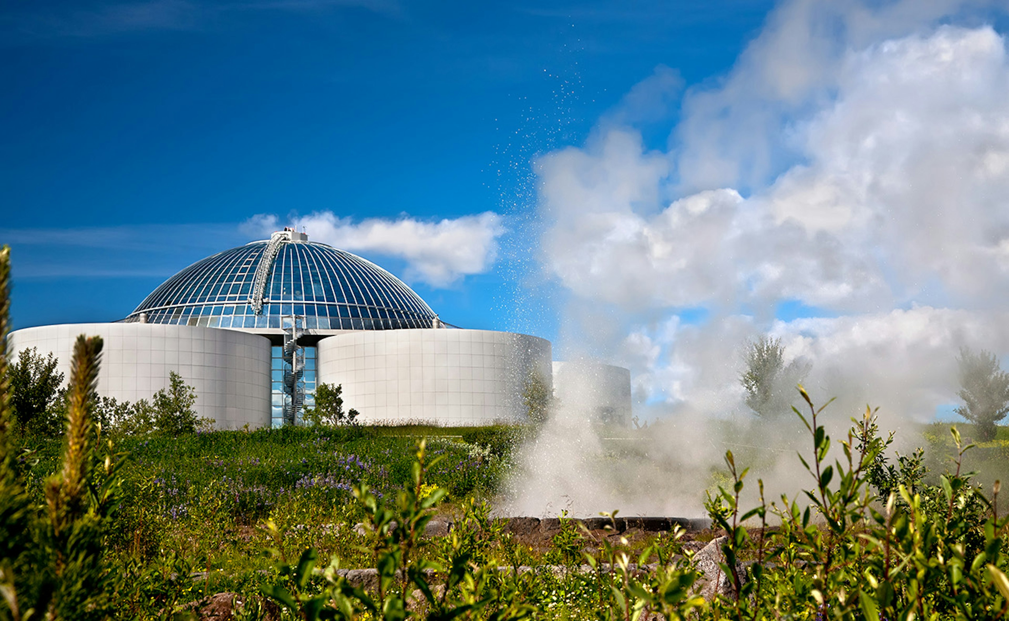 "Perlan" or "the Pearl" with a man-made geyser in front of it on a sunny day in Reykjavik