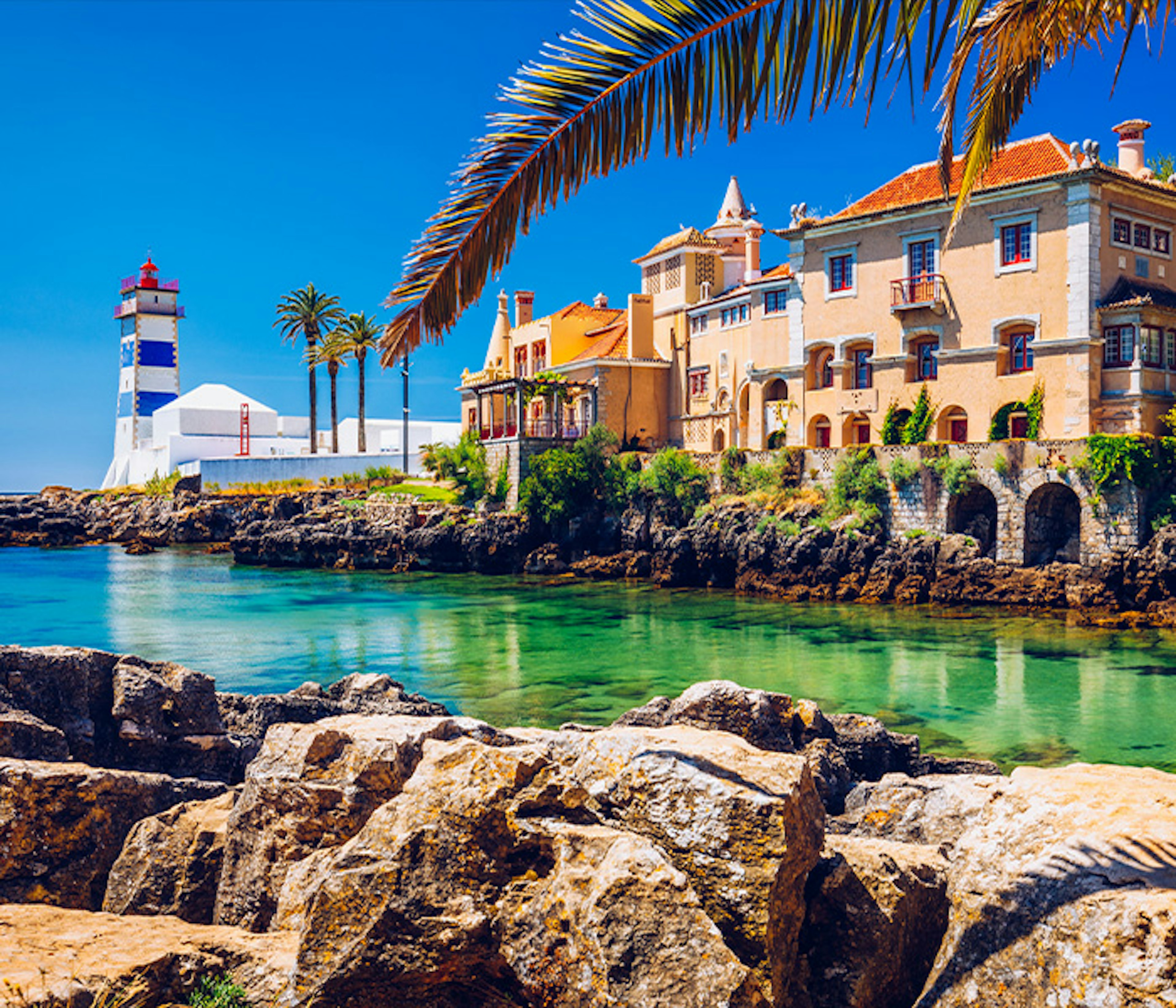 The clear sea in a small cove with a building and lighthouse on shore on a sunny day in Lisbon, Portugal