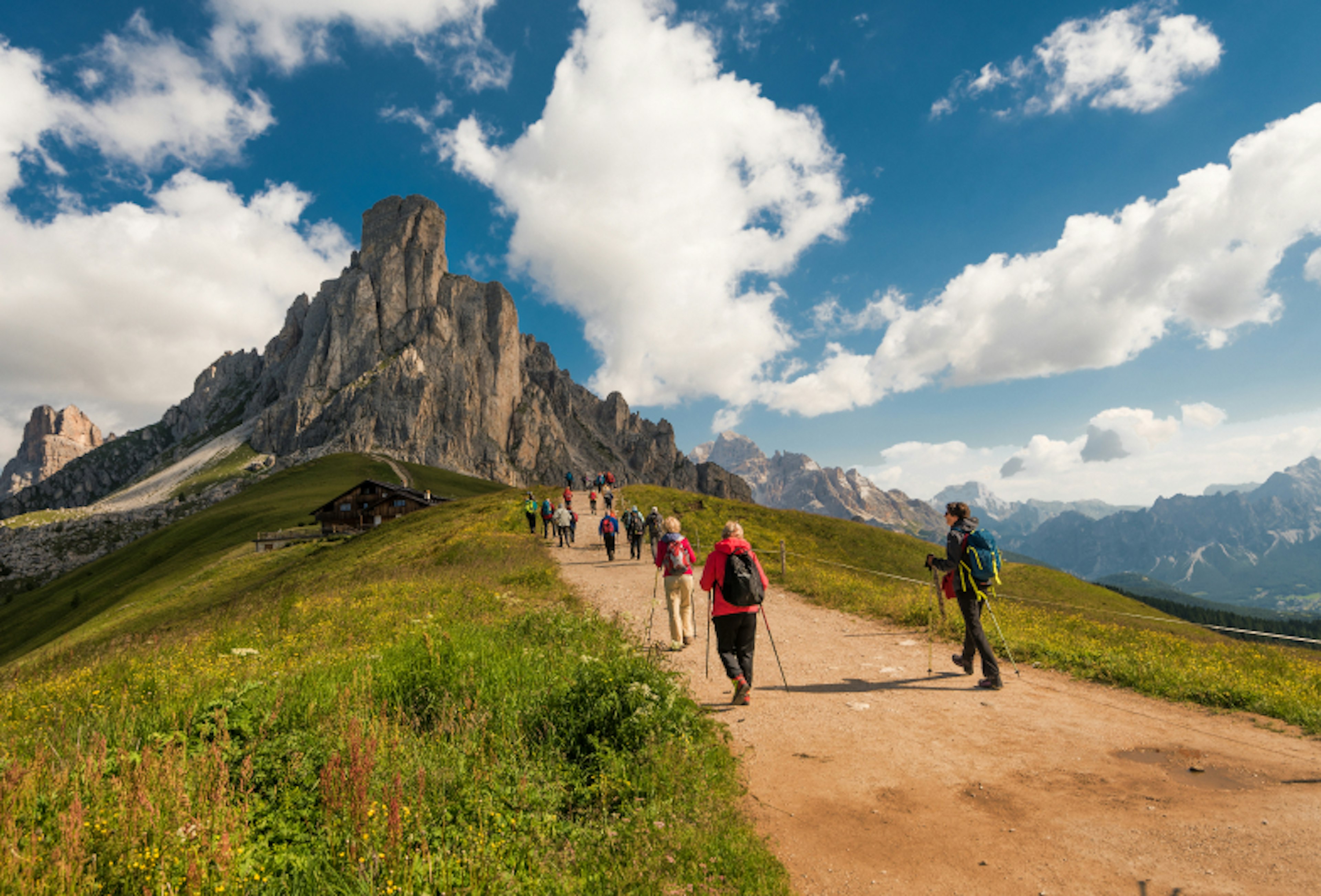 beautiful view of the Dolomites with hikers on path and grass in the foreground and hut, mountains and blue sky with clouds in the background taken on a sunny summer day