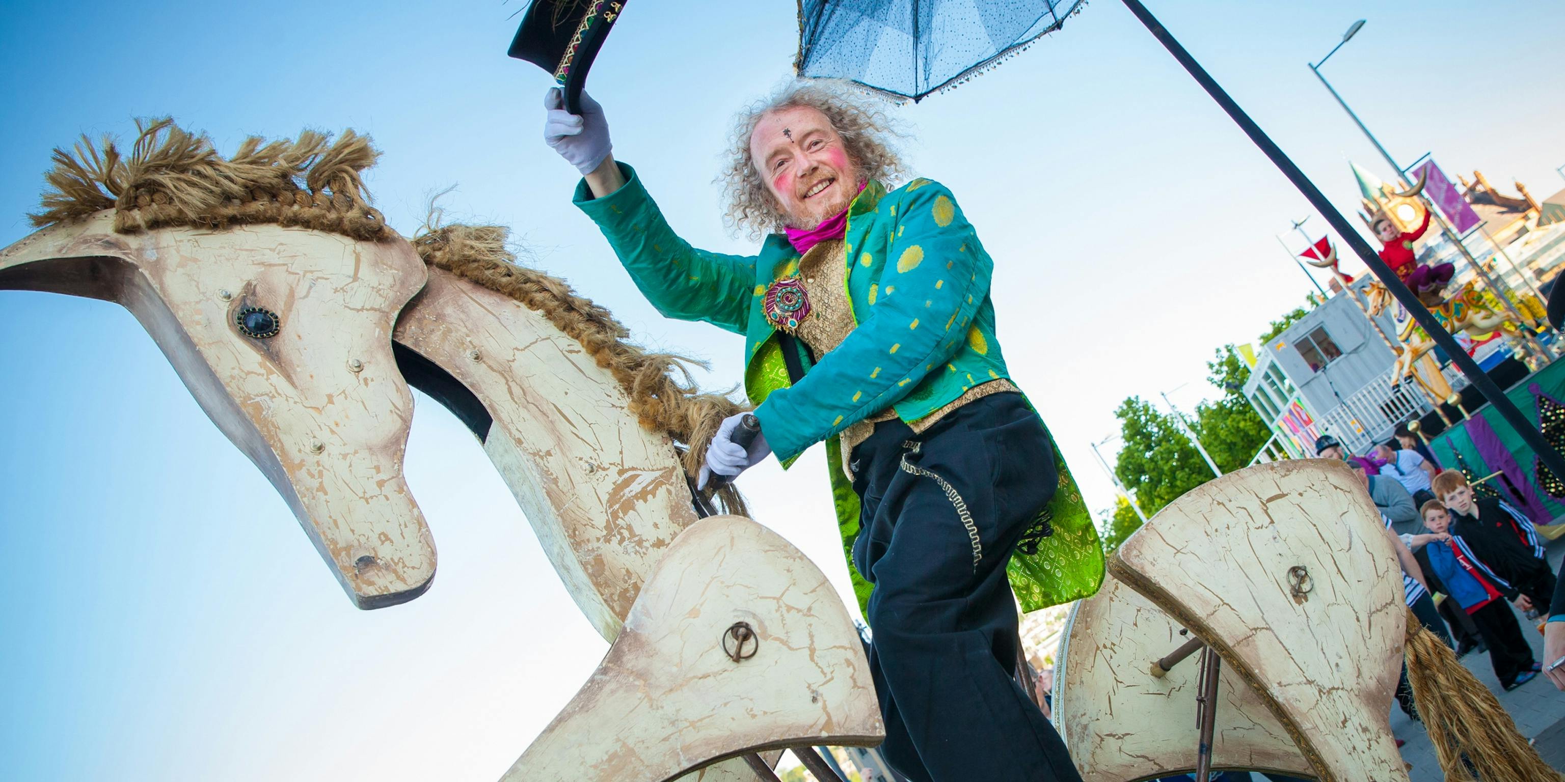 A performer in a green circus-style outfit sitting atop a wooden horse and lifting his hat