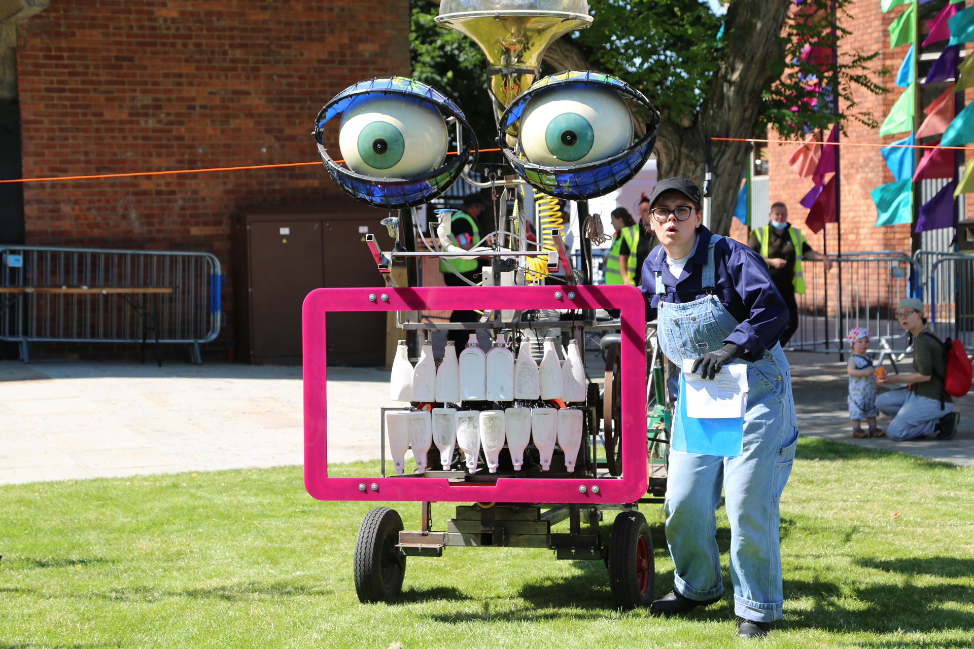 A performer in dungarees stands beside a large machine with bottles for teeth and large eyes