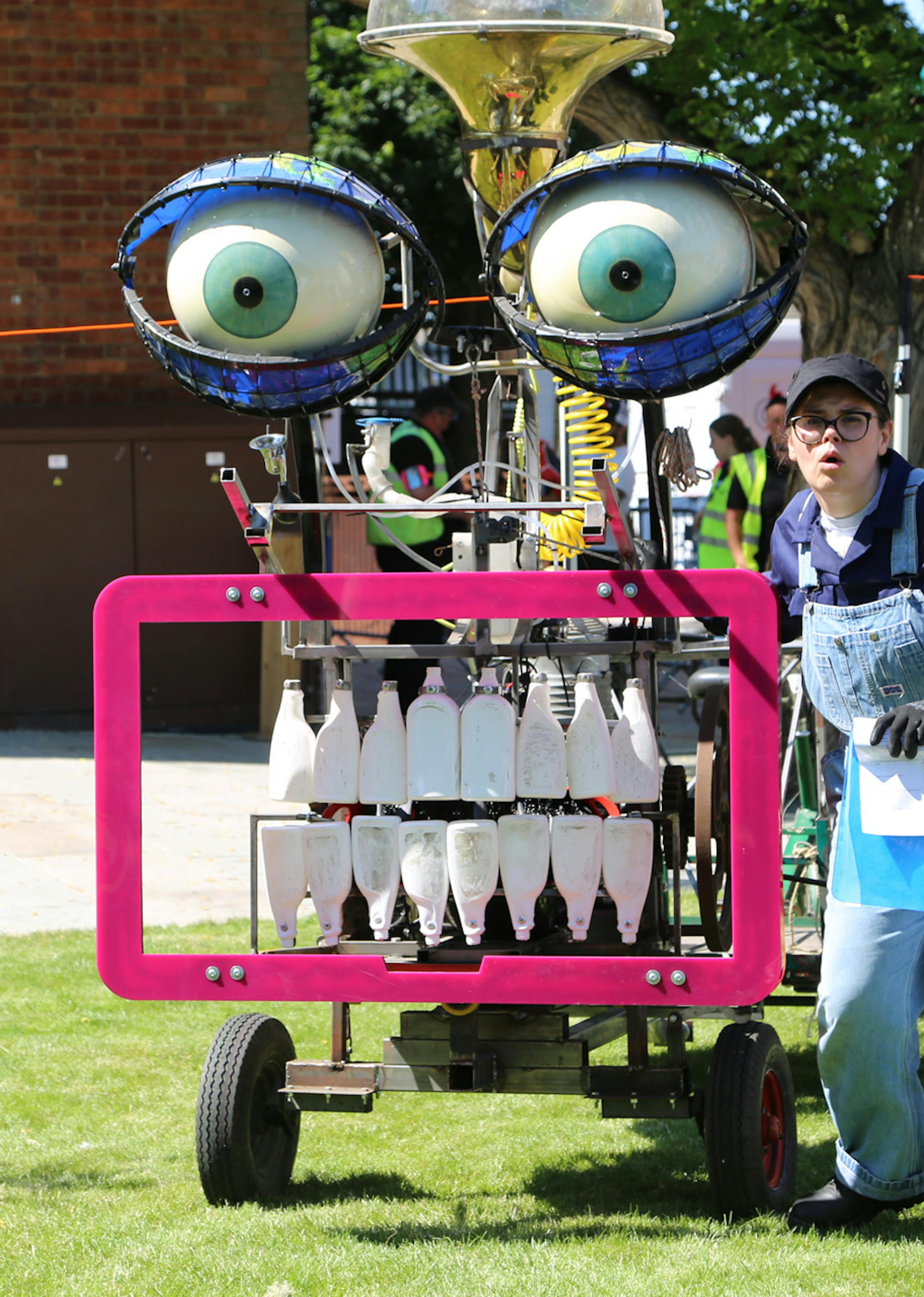A performer in dungarees stands beside a large machine with bottles for teeth and large eyes