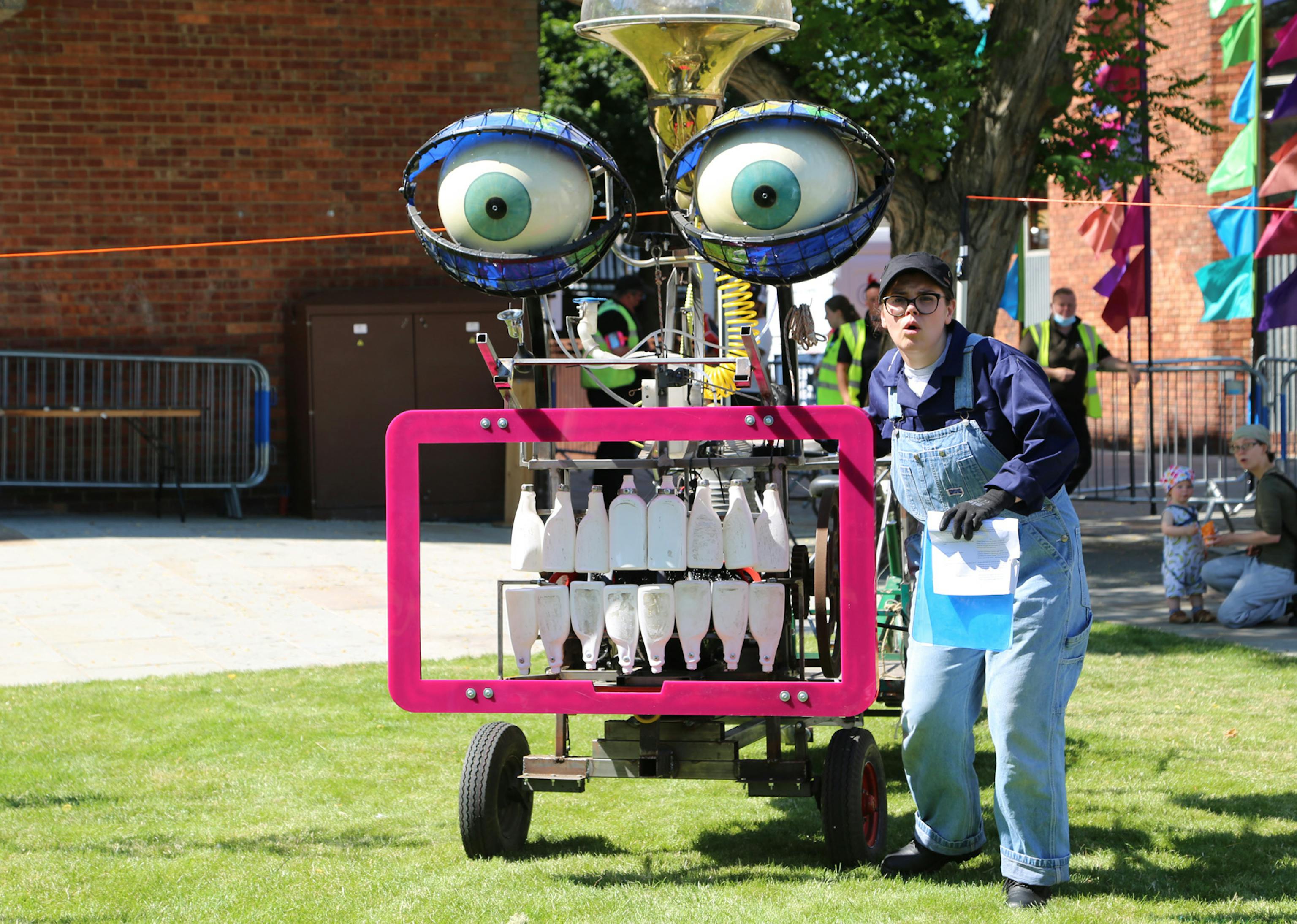 A performer in dungarees stands beside a large machine with bottles for teeth and large eyes