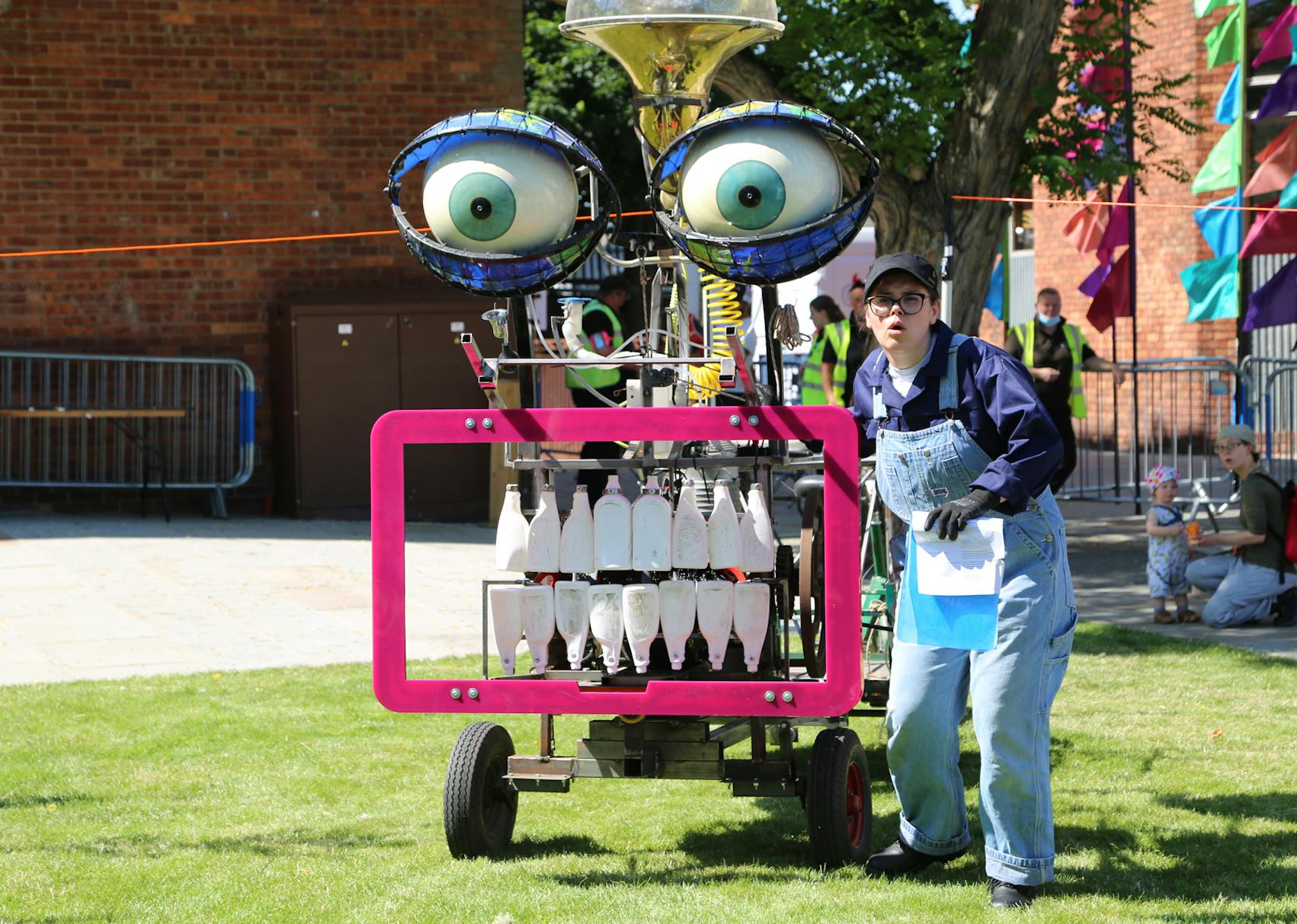 A performer in dungarees stands beside a large machine with bottles for teeth and large eyes