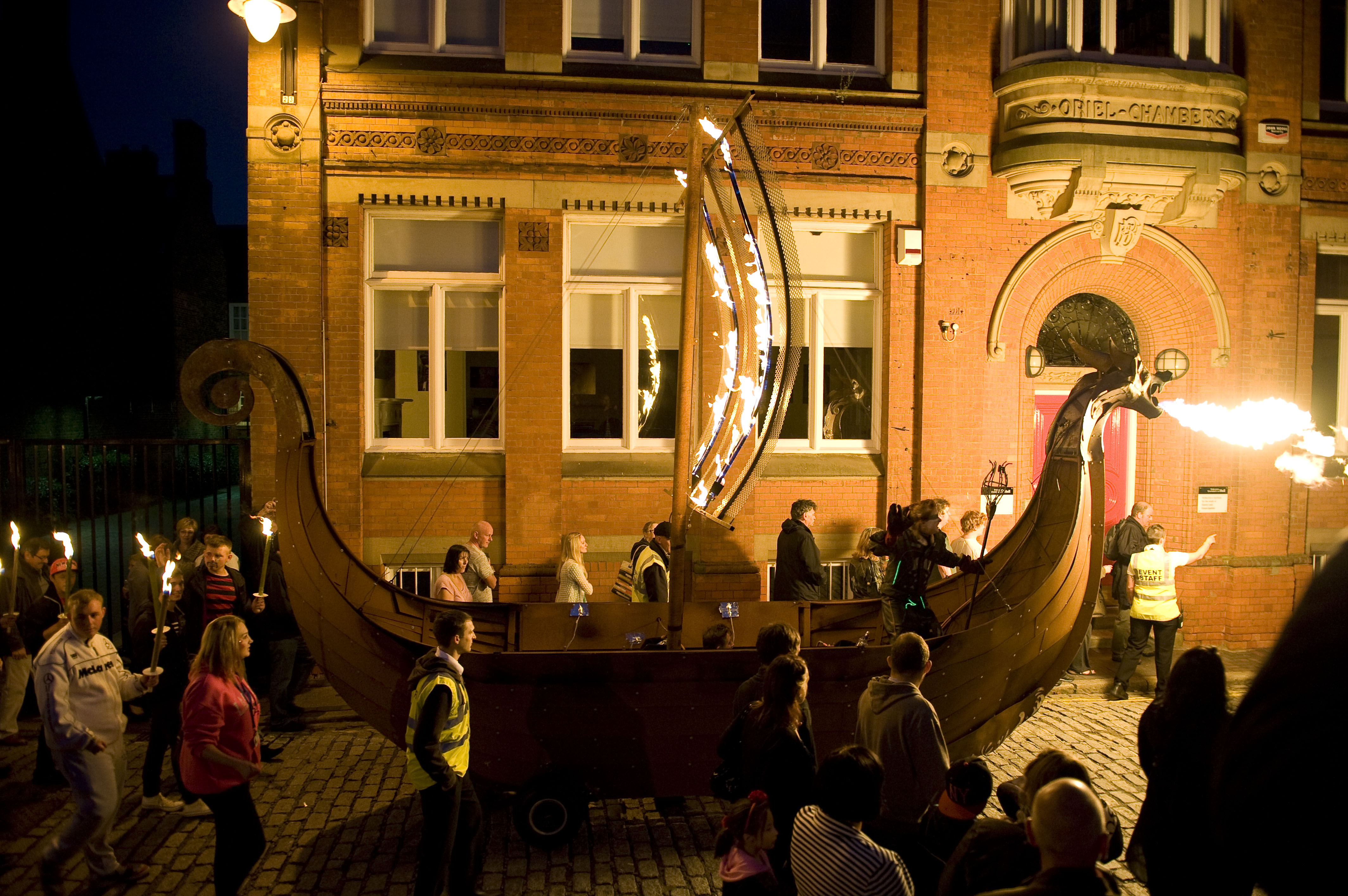 A wooden viking ship breathing fire, with fire in its sail being led through a town
